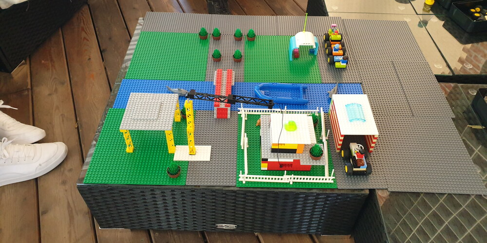 Die Legostadt steht auf einem niedrigen Tisch. Eine Brücke führt über einen Fluss, ein Haus mit Garage und Traktor, ein Kran und eine Bushaltestelle vervollständigen die Stadt.