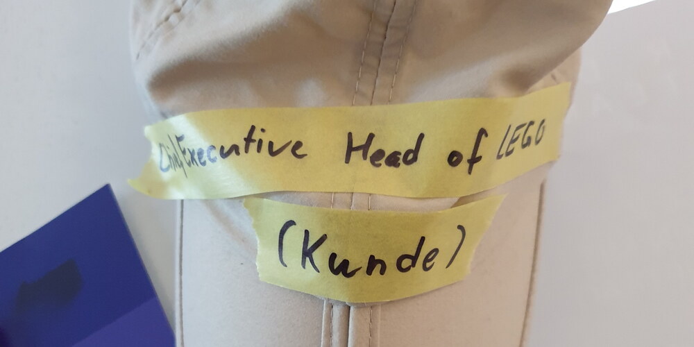 Eine helle Käppi mit beschriftetem Kreppband ("Chief Executive Head of LEGO") hängt an einem Whiteboard
